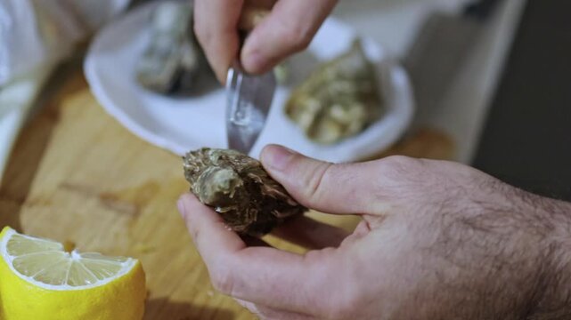 Man hand opens oysters at home on wooden surface using small knife. Lemon in frame, seafood preparation, kitchen scene, fresh ingredients, close view of shells and texture.