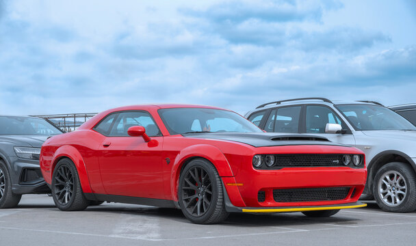 Red Dodge Challenger SRT Hellcat Widebody parked in a lot under a cloudy sky. A powerful American muscle car featuring a black hood and signature yellow splitter protector