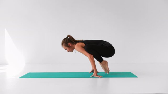 Flexible woman performing crow pose bakasana yoga asana on mat against white background in slow motion