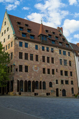 Naklejka premium Historic sandstone building in Nurnberg square under blue sky. Steep red roof and dormers frame a tower with a golden star. Arched floor and cobblestone plaza suggest medieval civic pride.