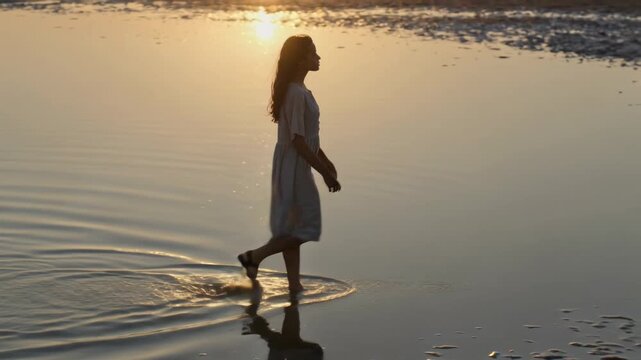 Walking woman wearing knee-length dress barefoot, passing shallow water creating ripples at sunset
