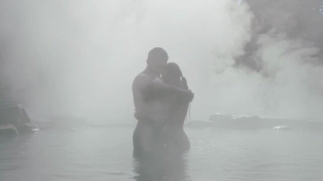 Reacting to cool air, swimwear-clad couple embracing in hot spring for warmth amid steam and rocks