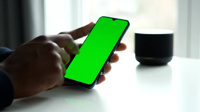 A person's hand interacts with a green screen smartphone on a white table near a smart speaker by a sunny window