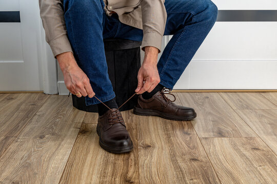 A man sitting on a black pouffe in a hallway puts on brown leather shoes, aches in his feet, and ties shoelaces
