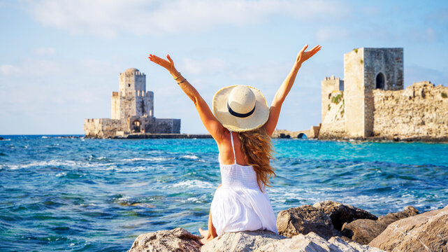 Young woman celebrating freedom and traveling with open arms overlooking the Venetian fortress and Bourtzi tower of Methoni, Greece.