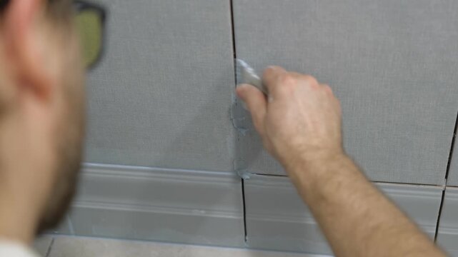 Worker using a rubber grout float to smooth grout between tiles. Carefully applying sealant to tiles joints. Workman hands smoothing caulk, using small tool to remove excess and create clean seam.