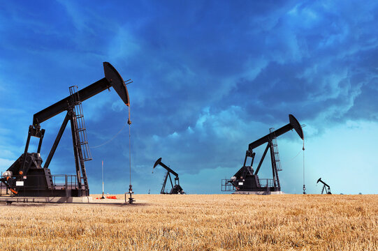Four traditional pump jacks stand silhouetted against a dramatic, stormy sky over a wide, golden-brown harvested field in a rural oil field landscape. High-resolution shot, cinematic and moody.