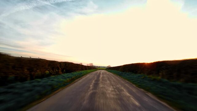 Fast driving along rural countryside road at sunset golden hour motion blur hedgerows point of view. Empty asphalt lane stretching into horizon warm evening sky travel freedom POV. Scenic road trip