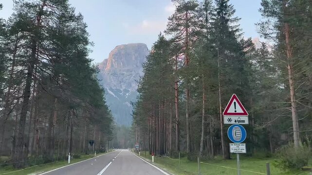  A scenic road in the Dolomites, Italy, showcases towering, jagged mountain peaks bathed in warm sunlight. The foreground includes a paved road lined with vehicles, bordered by sparse trees 