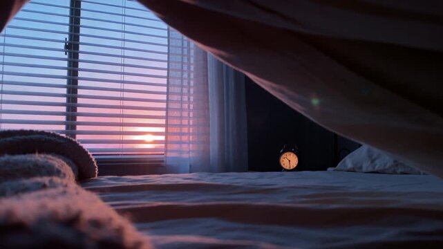 Rising sun casting warm light through blinds over bed, revealing textured throw, duvet, alarm clock