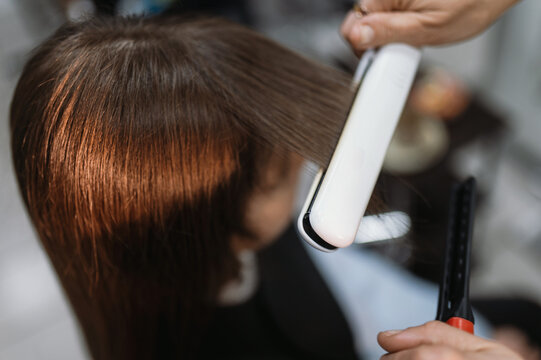 A hairdresser straightens and straightens a client's long, natural dark hair with a flat iron, curling iron, or styler at a salon. Professional hair styling and care at the hair salon.