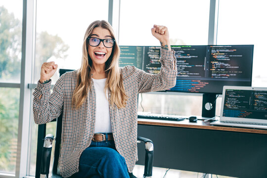 Female software developer celebrating a successful build at her desk with multiple monitors and laptop showing programming code in a modern home office