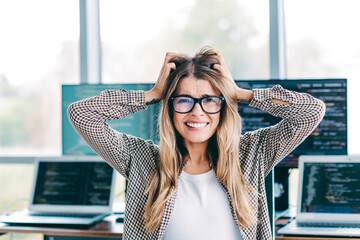 Frustrated female programmer pulling her hair in a modern office surrounded by laptops and...