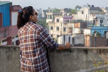 Side view of a Bengali woman wearing sunglasses and a plaid shirt, leaning on a concrete rooftop railing while looking at the city skyline. Ideal for urban lifestyle and travel themes © Sahadat