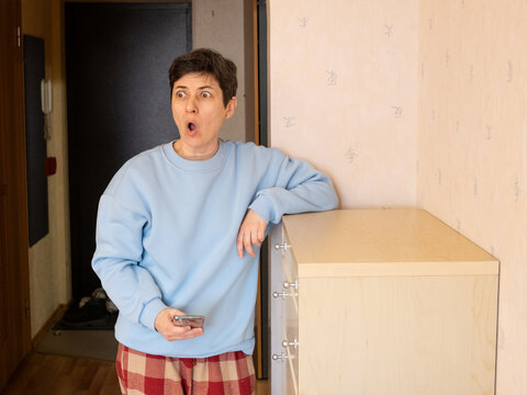 Middle aged woman reacting with surprise and shock while looking at smartphone screen in home interior, expressing unexpected reaction