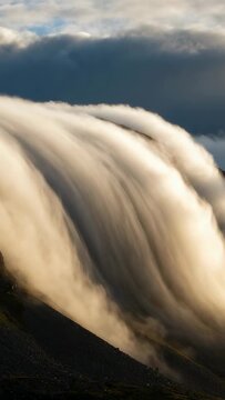 Vertical video: Spilling dense cloud mass flowing down steep mountain ridge, driven by cross wind