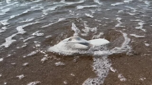A white flip-flop lay softly on the sandy beach as the calm ocean waves washed over it, creating a fine pattern of white foam.