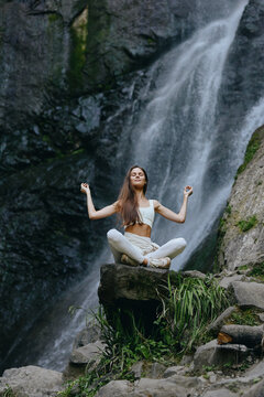Young woman meditating in white clothes sits cross-legged on rock near waterfall surrounded by lush greenery, expressing calmness and harmony in natural environment.