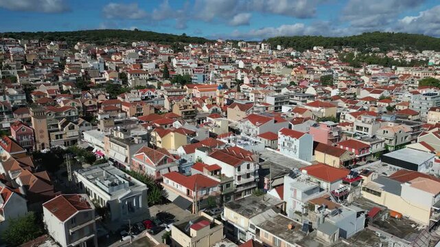 A densely packed town with colorful buildings and red-tiled roofs climbs a lush green hill under a bright blue, cloudy sky.