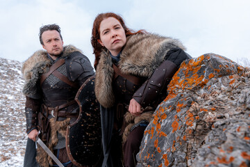 Naklejka premium Two medieval warriors, clad in furs and armor, pose stoically atop a rocky outcrop against a muted winter sky.