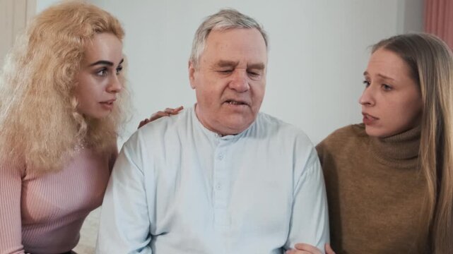 Loving daughters comforting sad elderly father, hugs him while sitting on the bed. Women caring older man at home. Family support, communication, relationships and mental health