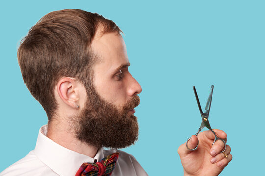 White man adult 30s with beard holding thinning scissors in side view in studio on blue background barber style. Concept of grooming service, haircut preparation and professional tool use