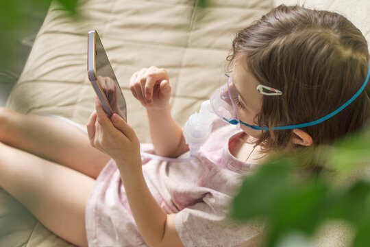 Child with nebulizer mask rests on sofa and looks at smartphone during home therapy, reflecting calm care and modern recovery.