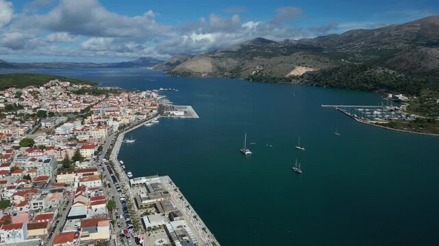 A picturesque coastal town with red-tiled roofs is bordered by a calm, blue bay dotted with sailboats and yachts. Mountains rise in the distance under a partly cloudy sky.