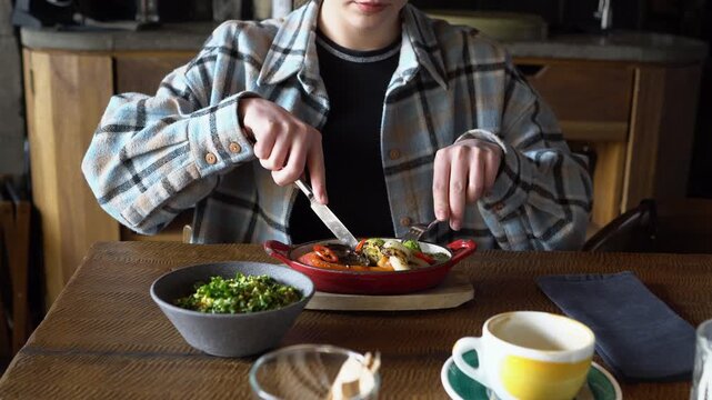 Young woman sitting in restaurant and eating grilled vegetables with fork and knife, cute girl having lunch in cafe