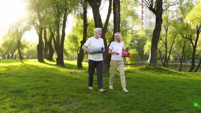 couple of elderly seniors go to yoga with mats and talk in the park, gray-haired pensioners walk after exercise and actively relax in the summer
