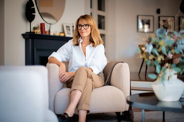 Obraz premium Blond haired smiling woman relaxing in an armchair in her modern home