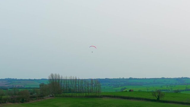 Paramotor pilot gliding above green rural hills farmland hedgerows and tree lines under wide cloudy sky. Extreme paramotoring flight drifting over countryside landscape outdoor aerial adventure sport