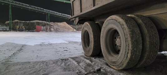 Close-up of heavy-duty semi-trailer wheels parked in a snow-covered industrial wood yard at night. Background features conveyors, log piles, and timber processing machinery under dim lighting. © DRONECYBORG
