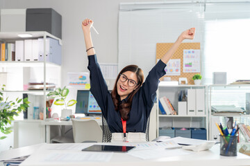 Successful Asian business woman stretching arms raised with joy sitting at office desk. Feeling happy after finish project achievement celebrate complete work career motivation productivity concept
