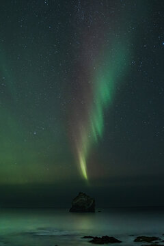 View of ethereal green and purple aurora borealis dances above the tranquil ocean, a lone rock formation silhouetted against the celestial display, Keflavik, Reykjanesbaer, Iceland.