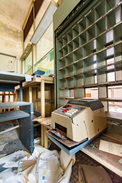 Abandoned retro accounting machine on a desk next to large mail sorting slots, ruined vintage post office or bank interior with scattered papers, lost place