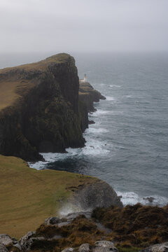 View of rugged cliffs meeting the turbulent sea under a misty sky, with the iconic Neist Point Lighthouse standing sentinel against the elements, Isle of Skye, Scotland.
