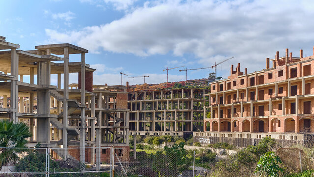 View of stark concrete skeletons of unfinished buildings rise against a blue sky dotted with puffy white clouds, a testament to halted construction, Manilva, Andalusia, Spain.