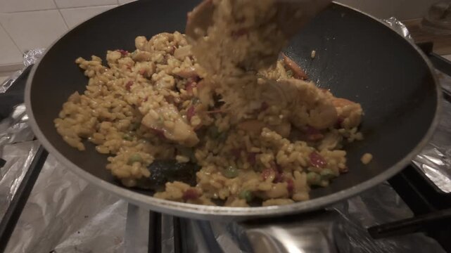 Close-up of seafood rice cooking in a frying pan on a kitchen stove. The dish includes shrimp, squid and rice, stirred with a wooden spoon. Concept of homemade cooking, traditional cuisine