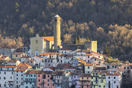View of pastel-hued houses cluster around a church tower, ancient stone ruins perched above, all bathed in the warm glow of a late afternoon, Caprigliola, Tuscany, Italy.