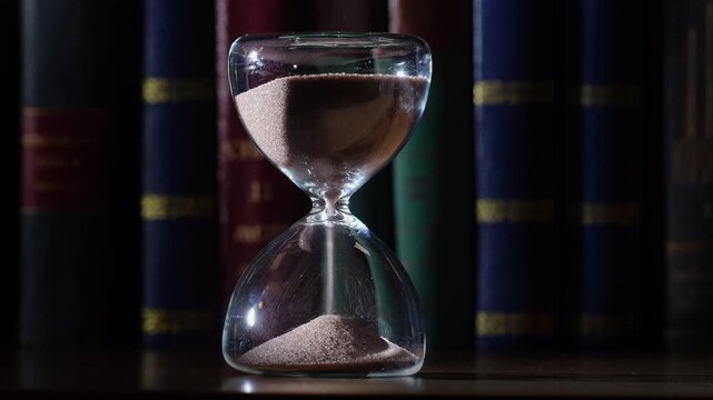 3 minute hourglass steady flow, bookshelf background. Shallow depth of field, gold embossed letters on old books in low light, centrally framed with copy space on both sides