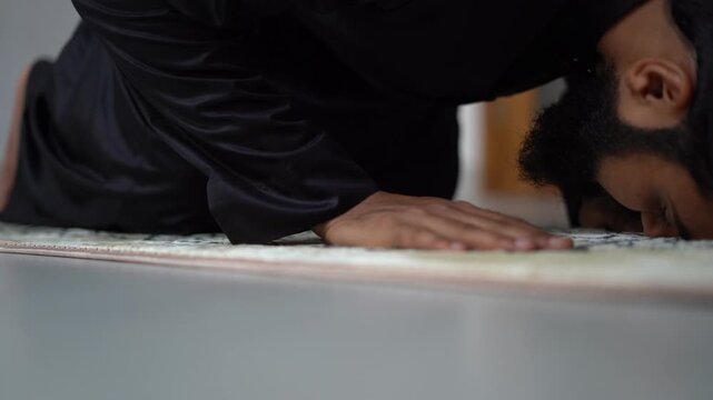 4K Close-up of Muslim man in black jubbah lowering to Sujud prostration on prayer mat, performing Salah, Islamic worship and devotion ritual.