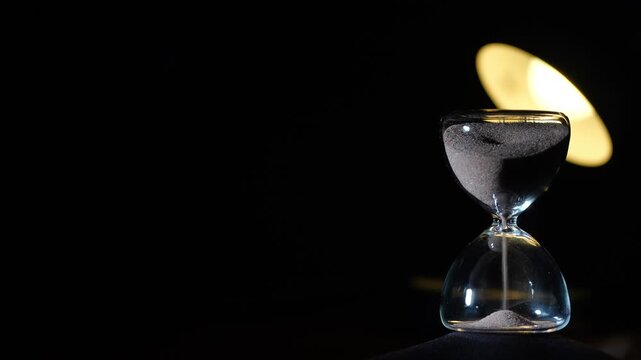 3 minute hourglass starting to flow on black background. Soft yellow reading lamp bokeh behind upper glass, right aligned with extreme copy space, cinematic low light, 3 min sand timer
