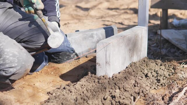 Mason Tapping Concrete Block Into Soil With Rubber Mallet, Closeup Hands Kneeling, Gloves, String Line Visible, Leveling Mortar Bed And Compacting Dirt, Outdoor Daylight Worksite