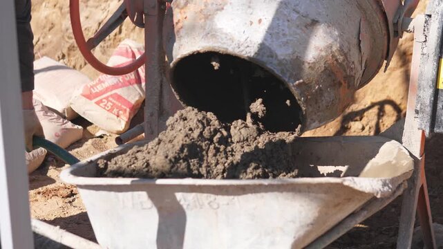 Concrete Pouring Underway. Construction Worker Carefully Pours Mixture From Drum Into Wheelbarrow. Laborer Tilts Mixer And Transfers Wet Concrete Into Wheelbarrow With Precision