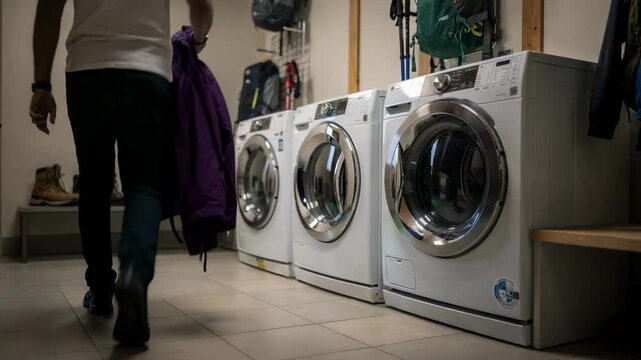 Medium shot of a guest laundry room featuring washing machines and dryers in focus with blurred hiking boots and gear racks in the background