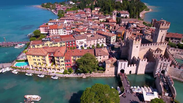 Sirmione 4k drone flyover of Scaliger Castle and bridge with people in Italy. Scenic aerial view of Garda lake peninsula and historic fortress architecture in Lombardy. Mediterranean travel scenery