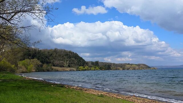 Scenic view of Lake Bolsena shoreline with green grass, trees and wavy blue water under a cloudy sky in Lazio, Italy
