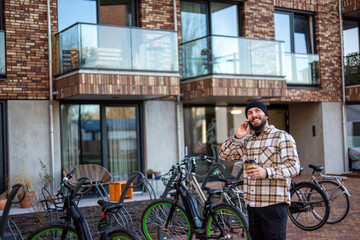 Obraz premium Young man talking on phone, holding coffee, smiling in urban street next to bicycles and modern building