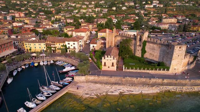 Italy Torri del Benaco aerial drone sunset over Lake Garda Scaliger Castle and small harbour with boats. Circular marina reveal with yachts and scenic water view Lombardy North Italy travel 4k